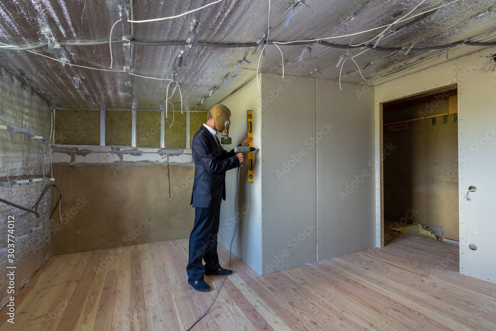 Fototapeta premium Strange man in businessman suit and gas protection mask inside a room under renovation works holding electric screwdriver and a level tools.
