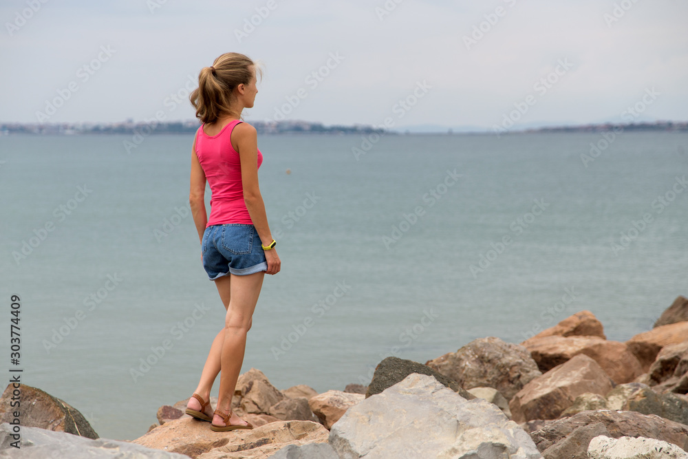 Young woman in summer clothes standing on big boulders on sea shore looking on horizon. Young woman in summer clothes standing on big boulders on sea shore looking on horizon.
