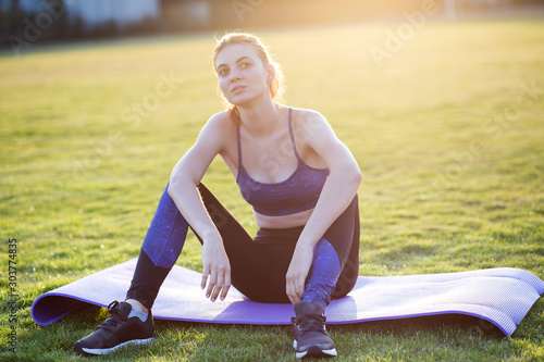 Young sportive woman in sports clothes sitting on training mat before doing exercises in field at sunrise.
