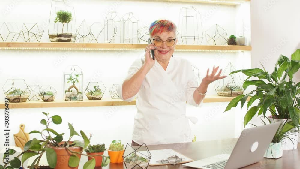 Emotional woman talking on mobile phone, explain varieties of plants. Laptop, paper, plants on table. Light room background Emotional woman talking on mobile phone, explain varieties of plants. Laptop, paper, plants on table. Light room background