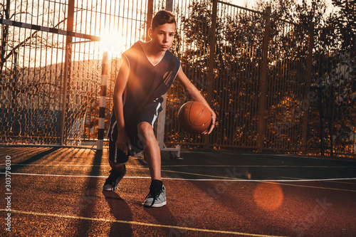 Sports and basketball. A young teenager in a black tracksuit is playing a basketball. Sunset light. Copy space © _KUBE_