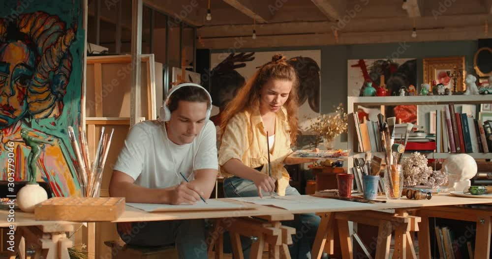Caucasian young couple of artists working in the cozy workshop, man listening to the music and drawing with a pencil and woman painting with paints. Caucasian young couple of artists working in the cozy workshop, man listening to the music and drawing with a pencil and woman painting with paints.