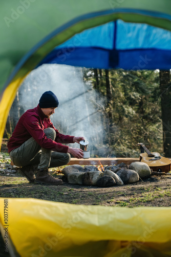 Image of a view from tent entrance of young man traveler pours coffee from coffee maker in metal cup with overlook amazing of forest. Explore travel real wilderness lifestyle