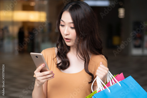 happy woman shopping using smartphone while holding shopping bag in shopping mall; concept of digital wallet technology, electronic payment, smartphone order, modern shopping technology