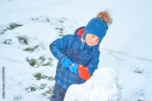 little boy in a hat and mittens makes a snowman in winter