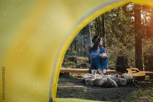 Calm shot from inside camping tent on a female traveller sitting near to campfire with overlook amazing views of forest, beautiful and inspiring camp location for outdoor vibes living lifestyle