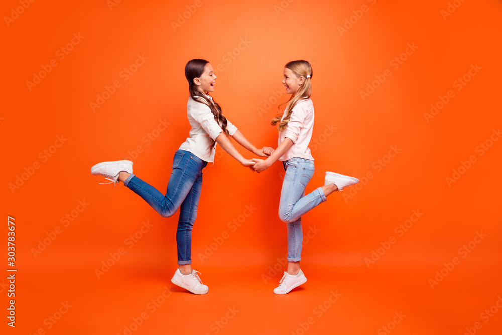 Full size profile side photo of positive cheerful two kids girls have free time hold hands rest relax wear modern clothing white sneakers isolated orange color background