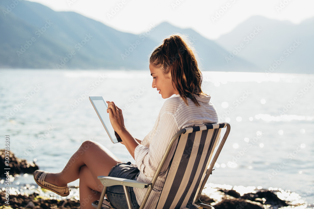 Smiling woman sitting on deck chair by the sea using tablet on a sunny day Smiling woman sitting on deck chair by the sea using tablet on a sunny day