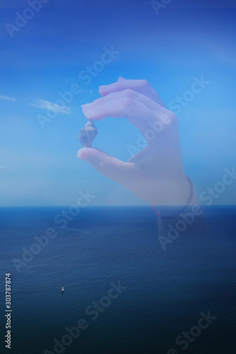 Fototapeta Naklejka Na Ścianę i Meble -  Silhouette of a hand holding a shell on a background of the sea and a sailboat