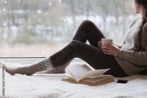 Thoughtful young brunette woman with morning cup of coffee looking through the window, blurry winter outside. 