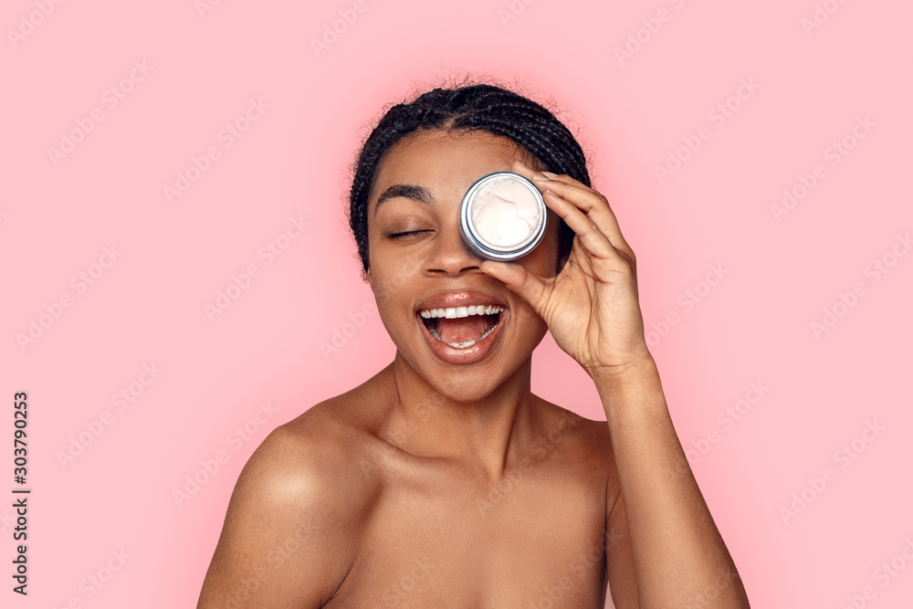 Beauty Concept. Young african woman isolated on pink covering eye with cream jar laughing cheerful - obrazy, fototapety, plakaty 