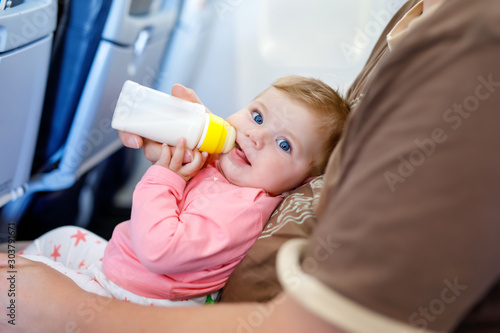 Father holding baby daughter during flight on airplane going on vacations. Baby girl drinking formula milk from bottle. Air travel with baby, child and family concept. Tired man traveling with kids.