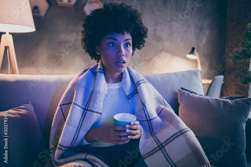 Unbelievable series concept. Astonished afro american girl sit couch watch film hold cup coffee look voiceless covered with checkered plaid blanket in house evening indoors photo