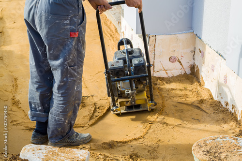 worker compresses sand in blind area around building with special working tool (tamping machine or vibratory plate)