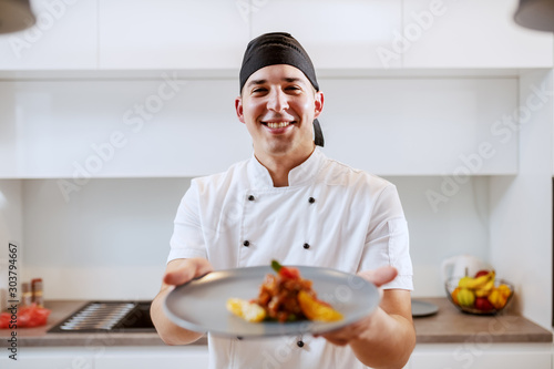 Portrait of Caucasian chef in uniform holding plate with salmon and orange fruit. Selective focus on chef.