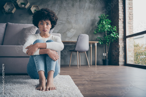 Photo of pretty dark skin curly lady sitting floor near sofa sad eyes full of fear hold knees near chest afraid to be alone home wear casual sweater jeans outfit flat indoors
