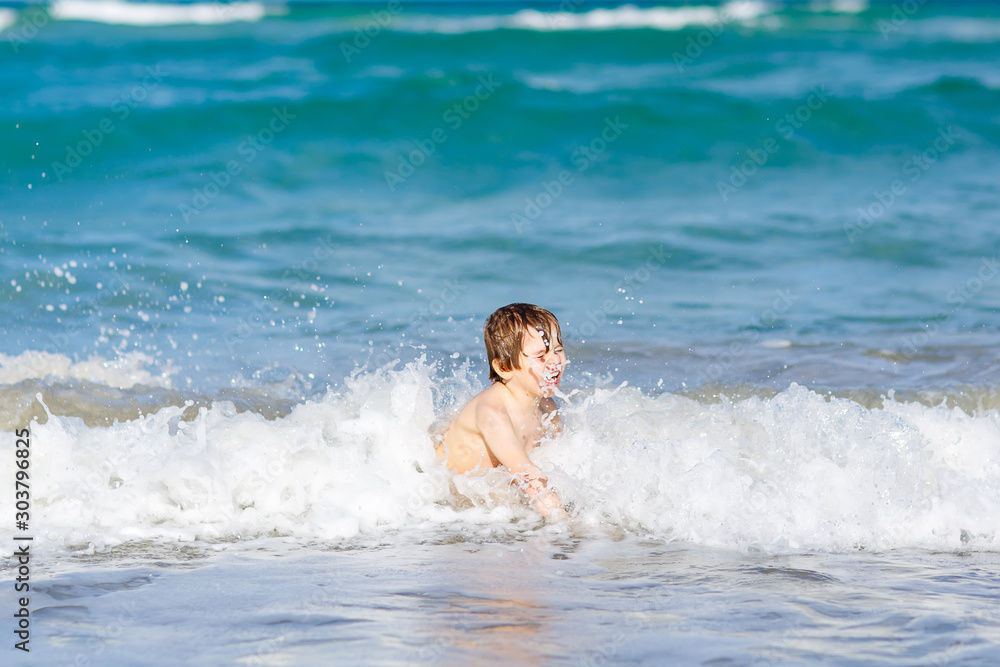 Adorable little blond kid boy having fun on ocean beach. Excited child playing with waves, swimming, splashing and happy about family vacations in Miami, Florida, USA..
