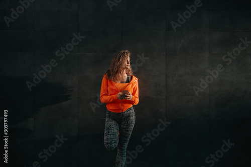 Charming smiling caucasian woman in sportswear leaning on black wall and using smart phone while resting after exercises.