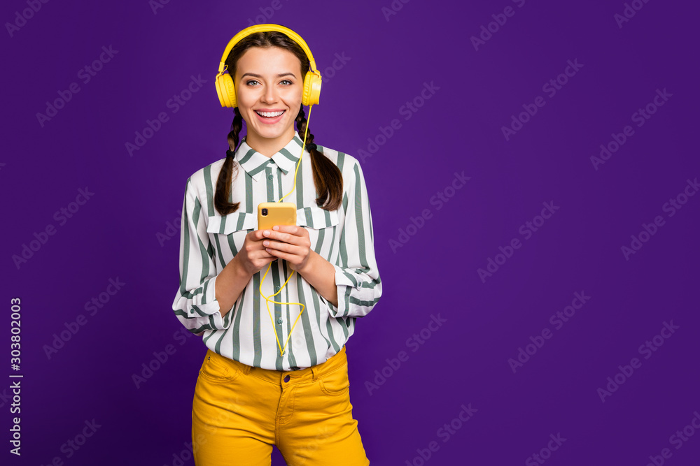 Photo of beautiful youngster lady holding telephone positive vacation time listen music wear cool earflaps striped shirt yellow trousers isolated purple color background Photo of beautiful youngster lady holding telephone positive vacation time listen music wear cool earflaps striped shirt yellow trousers isolated purple color background