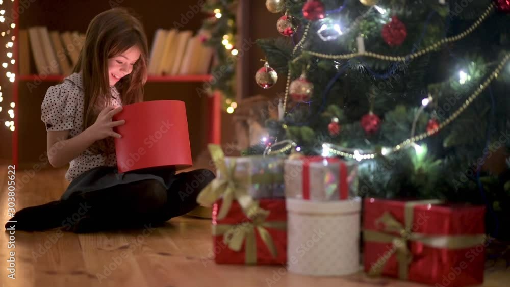 Cute girl sitting in the evening on the floor in the room opens a gift box and smiles. Against the background of the Christmas tree and festoon garlands. Christmas and New Year. Holidays. Cute girl sitting in the evening on the floor in the room opens a gift box and smiles. Against the background of the Christmas tree and festoon garlands. Christmas and New Year. Holidays.