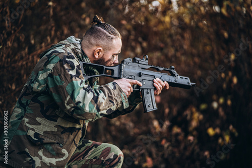 Portrait of a camouflage soldier with rifle and painted face playing airsoft outdoors in the forest. Side view