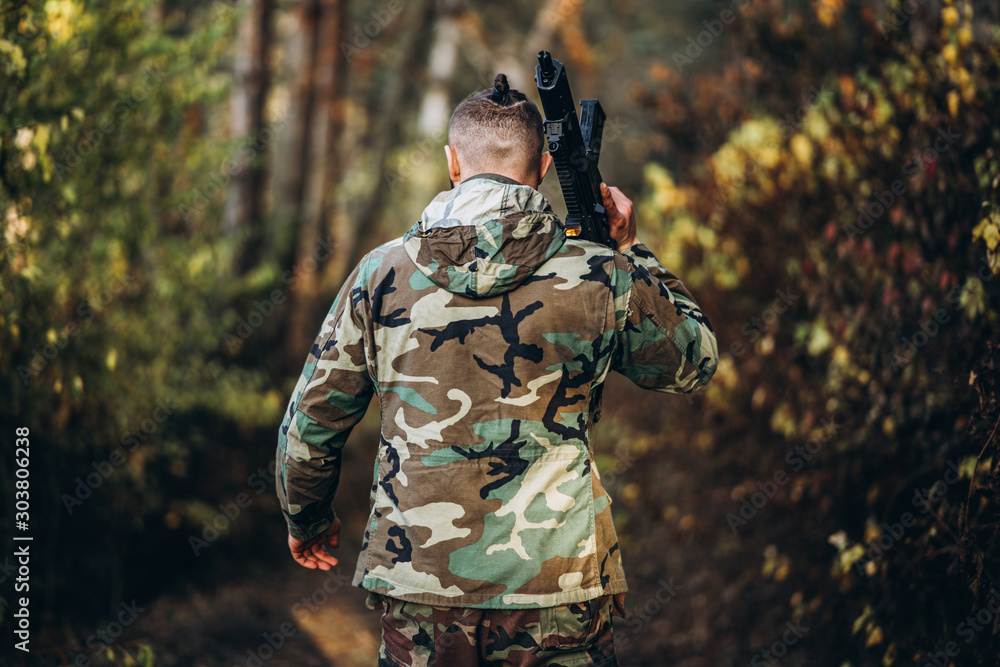 A soldier in camouflage uniform with a rifle on his shoulder walk in the forest. Back view. Airsoft