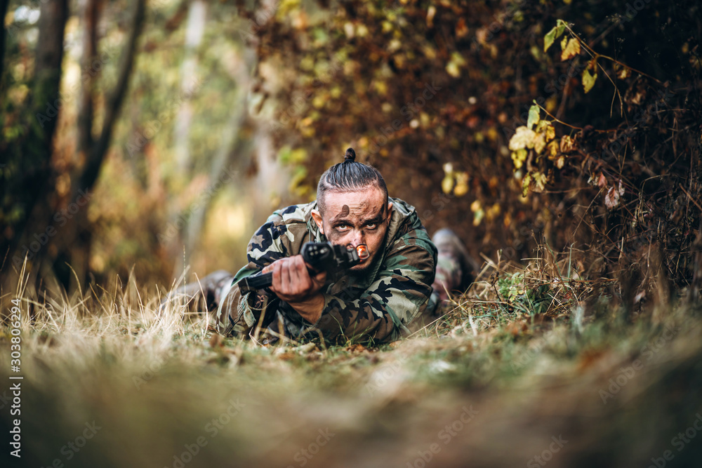 Φωτογραφία Portrait of a camouflage soldier with rifle and painted face lying in the grass aiming at the rifle