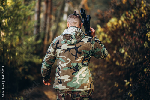 A soldier in camouflage uniform with a rifle on his shoulder walk in the forest. Back view. Airsoft