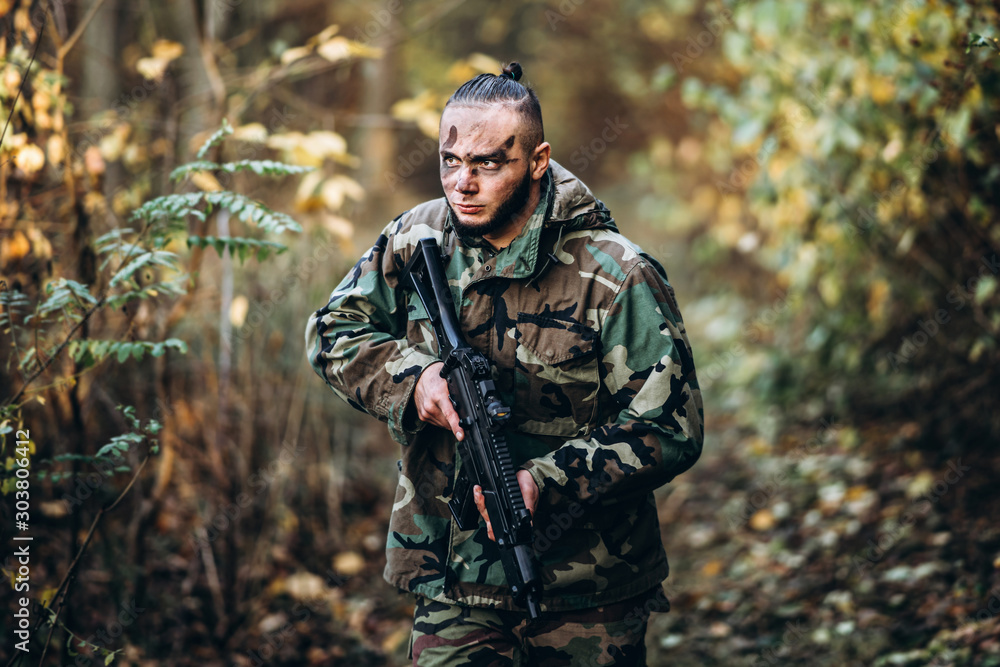 Portrait of a camouflage soldier with rifle and painted face playing airsoft outdoors in the forest