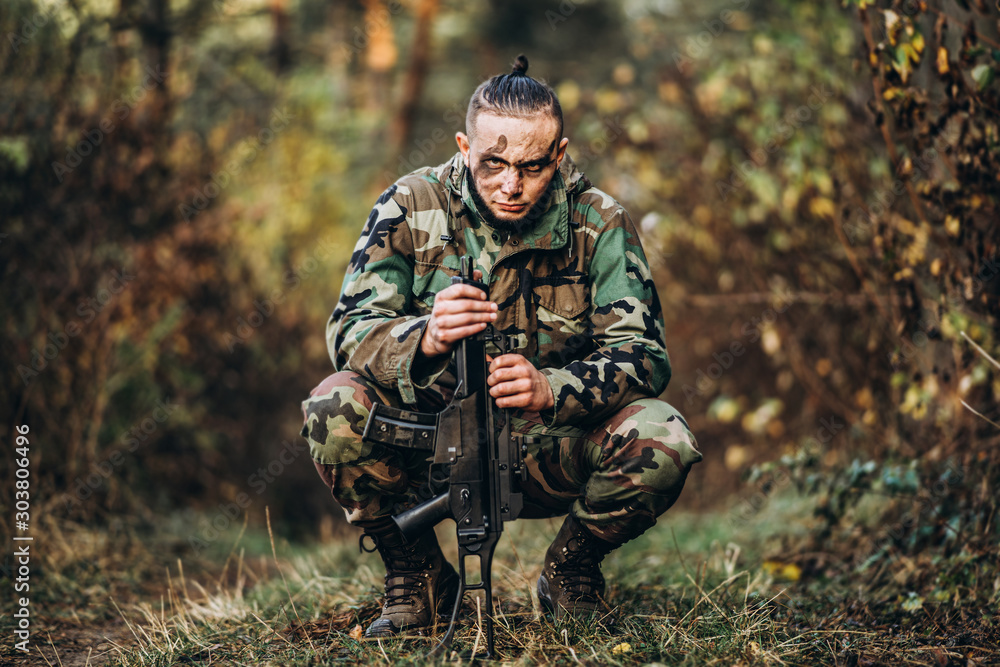 Portrait of a camouflage soldier with rifle and painted face sitting in the grass. Airsoft outdoors in the forest