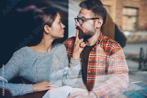 Angry young woman making him stop talking during quarrel.Hipster girl told secret to boyfriend inn and showing shh sitting in coworking space.Bearded man under pressure of girlfriend photo