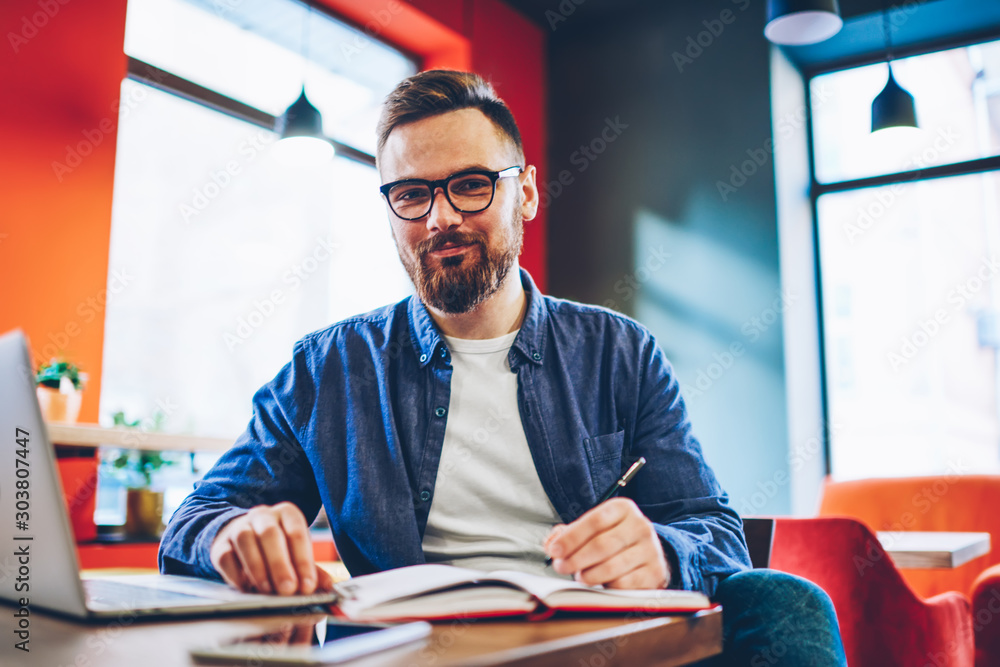 Obraz na plátně Half length portrait of successful bearded student doing homework and studying at laptop computer while smiling at camera