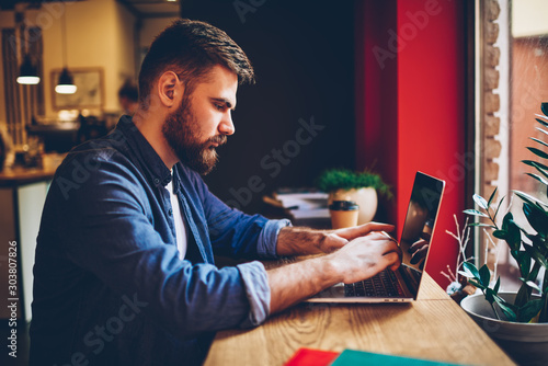 Side view of bearded freelancer keyboarding text information on modern laptop computer connected to wireless 4G internet.Male graphic designer working remotely at netbook in stylish coworking space