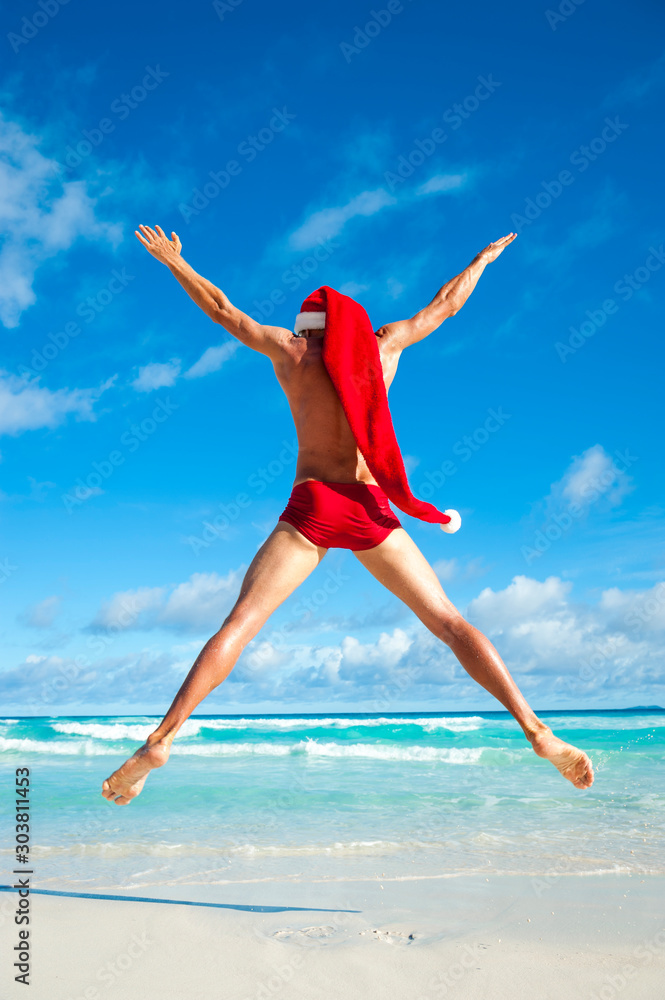 Excited Santa with extra long hat jumping in skimpy red speedos on the shore of a beach in a tropical holiday celebration