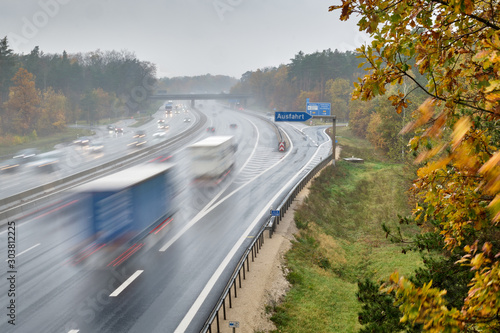 The highway A3 near the exit " Nürnberg Nord " (in English: Nuremberg North ) with many cars with lights switched on driving with speed on a wet rainy day in November with autumn forest around