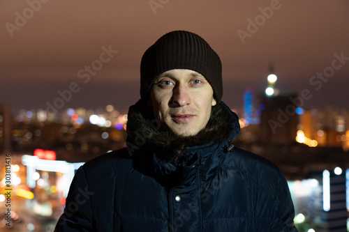 street portrait of a young unshaven European man with gray eyes wearing a knitted warm hat and a warm winter jacket, standing at night on the roof of the house, posing against the background of the ni