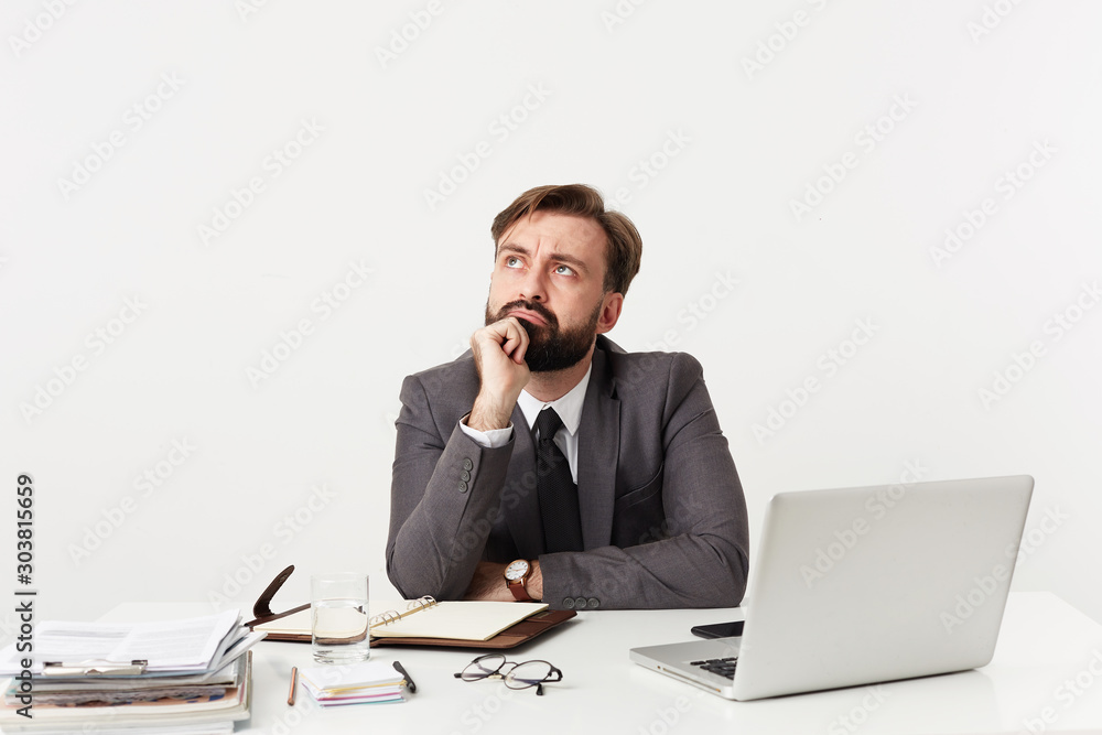 Puzzled young bearded brunette man with short haircut wearing formal clothes and wristlet watch while sitting at table with modern laptop and working notes over white background Puzzled young bearded brunette man with short haircut wearing formal clothes and wristlet watch while sitting at table with modern laptop and working notes over white background