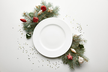 Top view of festive Christmas table setting on white background with decorated pine branch, plate and confetti