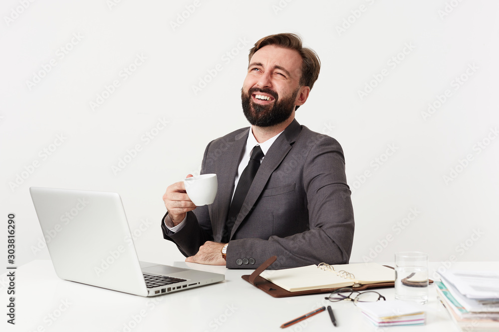 Indoor photo of joyful young bearded brunette man working in office with his laptop, laughing happily while drinking tea, wearing grey suit and tie over white background Indoor photo of joyful young bearded brunette man working in office with his laptop, laughing happily while drinking tea, wearing grey suit and tie over white background