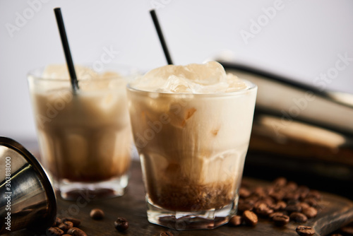 selective focus of white russian cocktail in glasses with straws on wooden board with coffee grains isolated on grey