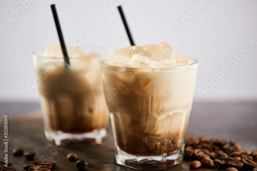 selective focus of white russian cocktail in glasses with straws on wooden board with coffee grains isolated on grey