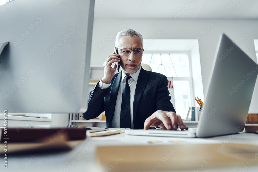 Senior man in elegant business suit talking on the phone while working in modern office Senior man in elegant business suit talking on the phone while working in modern office