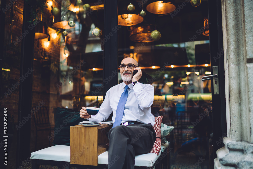 Senior man in glasses sitting and talking on phone against glass Senior man in glasses sitting and talking on phone against glass