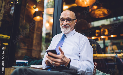 Senior smiling man in glasses texting on smartphone against glass