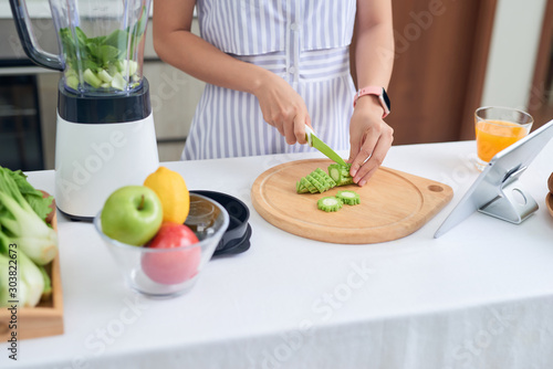 Woman cutting fruit vs vegetable to make smoothies at the kitchen from recipe on tablet.