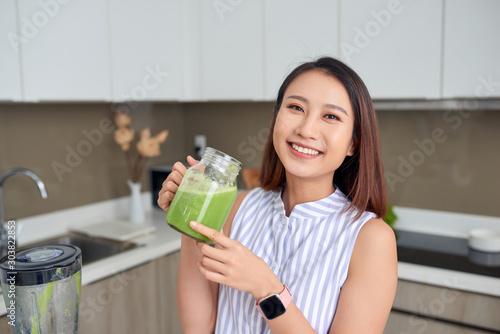 Smiling young Asian woman holding green smoothie in the kitchen at home.