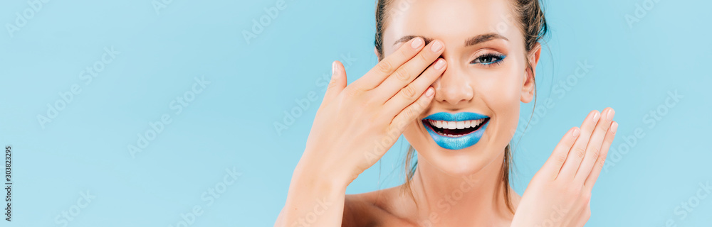 Obraz premium happy beautiful woman with blue lips covering eye with hand isolated on blue, panoramic shot