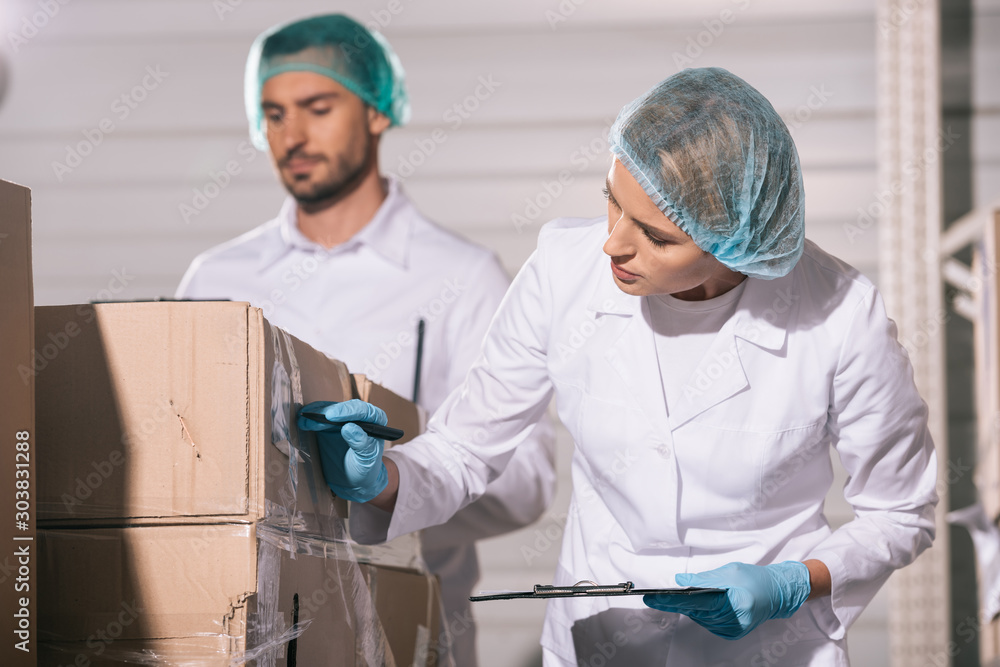 selective focus of attractive storekeeper writing on cardboard box near handsome colleague selective focus of attractive storekeeper writing on cardboard box near handsome colleague