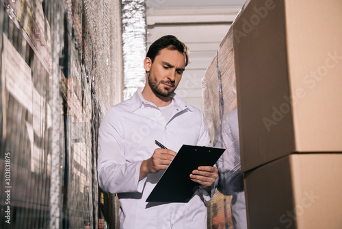 concentrated storekeeper in white coat writing on clipboard in warehouse photo
