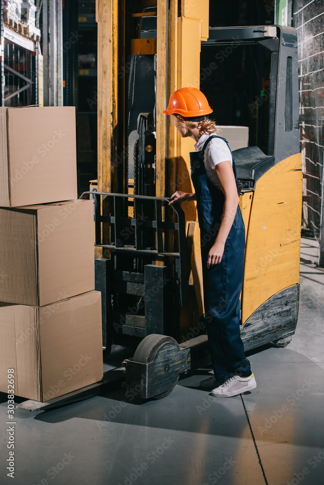 workwoman in overalls and helmet standing near foklift loader in warehouse workwoman in overalls and helmet standing near foklift loader in warehouse